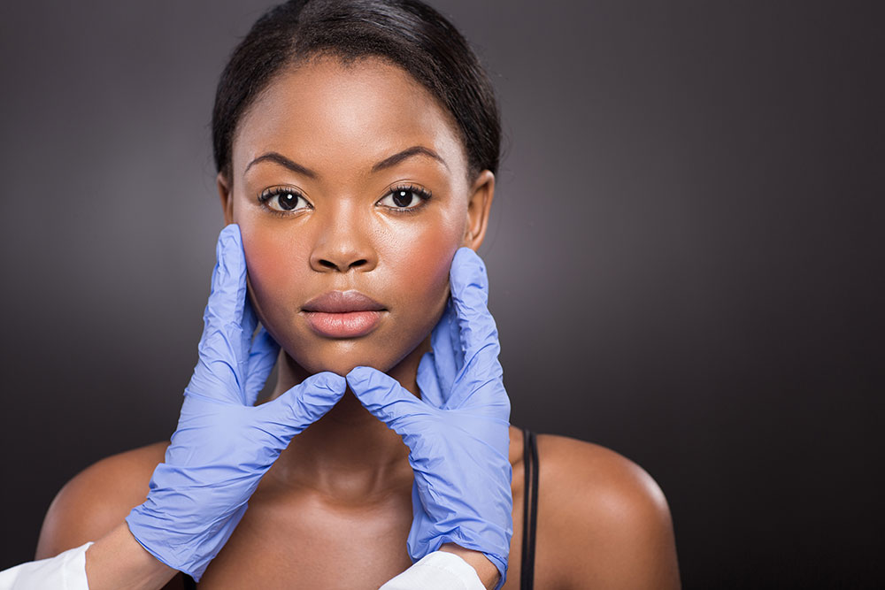 Médico com luvas roxas segurando rosto de mulher negra para ilustrar conteúdo sobre cirurgia plástica reconstrutora.